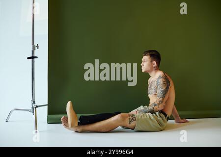 Ein junger Mann sitzt nachdenklich in einem Studio und zeigt seine Tattoos und seine einzigartige Beinprothese vor einem modernen Hintergrund. Stockfoto