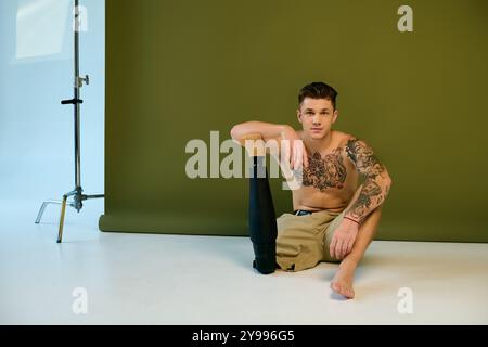 Ein tätowierter junger Mann sitzt auf dem Boden in einem Studio und zeigt stolz seine Beinprothese und seine komplizierten Tätowierungen. Stockfoto