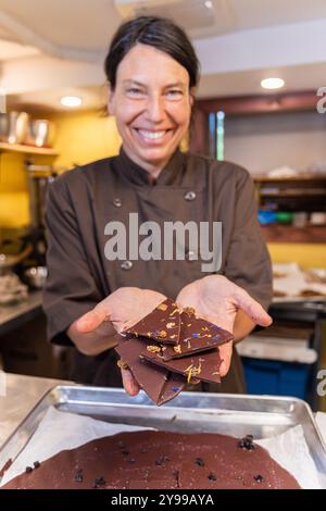 Chocolatierin lächelt und hält Stücke dunkler Schokolade mit Nüssen und Blumen in der Hand. Selektiver Fokus Stockfoto