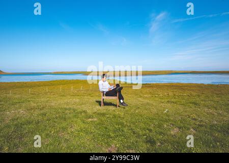 Ein Mann sitzt auf einem grünen Feld neben einem Holzstuhl, mit einem ruhigen Fluss und blauem Himmel im Hintergrund. Die friedliche Szene spielt im Stockfoto