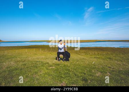 Der Mann sitzt auf einem hölzernen Stuhl in einem riesigen grünen Feld und macht mit seinem Handy ein Selfie vor dem Hintergrund eines ruhigen Flusses und blauen Himmels im Westen Islands. Stockfoto