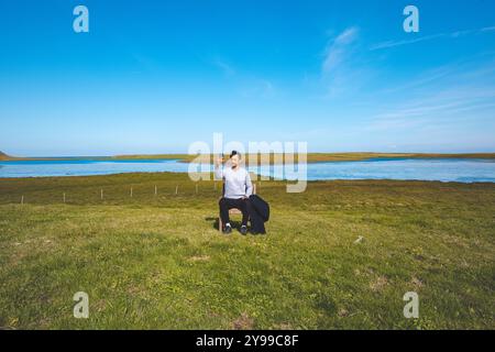 Der Mann sitzt auf einem hölzernen Stuhl in einem riesigen grünen Feld und macht mit seinem Handy ein Selfie vor dem Hintergrund eines ruhigen Flusses und blauen Himmels im Westen Islands. Stockfoto