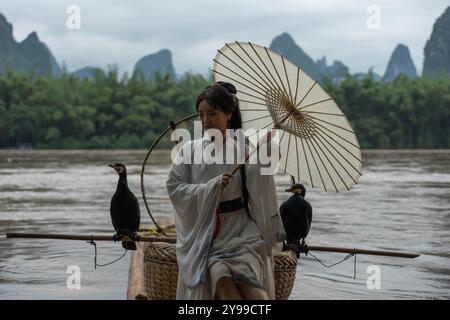 Hanfu Girl sitzt auf einem Bambusfloß und hält einen Regenschirm, während sie ihre Haare fixiert Stockfoto