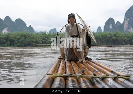 Hanfu Girl mit einem Schirm posiert auf Bambusfloß auf dem Fluss mit zwei Kormoranen. Xingping, China Stockfoto