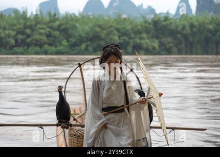 Chinesisches Hanfu-Mädchen mit einem Schirm, der Arme auf einem Bambusfloß im Fluss Li öffnet. Vertikal Stockfoto