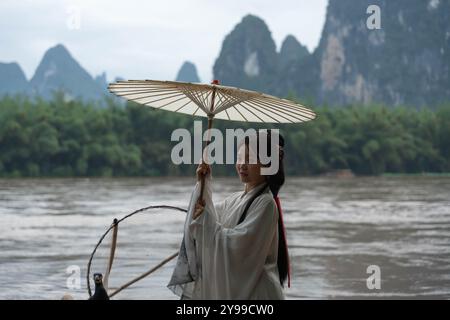 Wunderschönes Hanfu-Mädchen mit Regenschirm, der auf einem Bambusfloß steht, Arme ausstreckt und ihr Haar fixiert Stockfoto