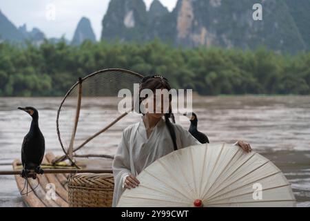 Hanfu Girl mit einem Schirm posiert auf Bambusfloß auf dem Fluss mit zwei Kormoranen. Xingping, China Stockfoto