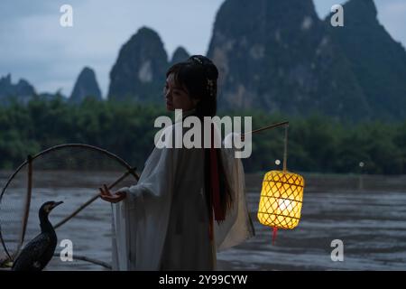 Hanfu Girl hält eine beleuchtete Laterne auf einem Bambusfloß mit Kormoranvögeln in der Nacht Stockfoto