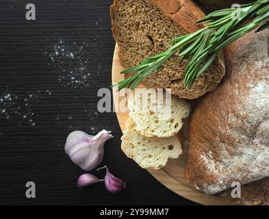 Frisch gebackenes Brot und Knoblauch auf Holzteller mit Rosmarin auf dunklem Hintergrund. Menü- und Café-Design Stockfoto