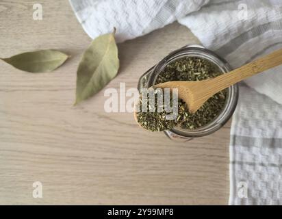Glasbehälter mit getrockneten provenzalischen Kräutern und einem Holzlöffel auf einem Tisch mit Lorbeerblatt in gemütlicher Küche. Getrocknete Rosmarin-Gewürze Stockfoto