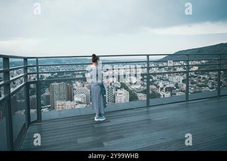 Eine Frau in legeren Kleidern blickt über eine weitläufige Stadtlandschaft und genießt den Blick von einer hohen Aussichtsplattform, während Wolken über ihnen Rollen. Stockfoto
