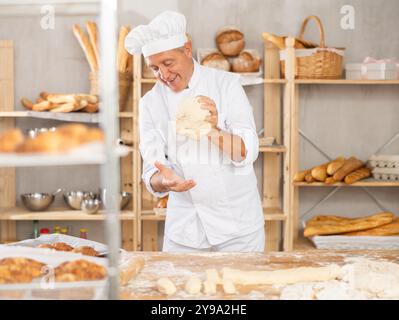 Ein älterer Bäcker steht an seiner Werkbank und kntet und formt Teig, um Brot, Croissants und Baguettes zu machen Stockfoto