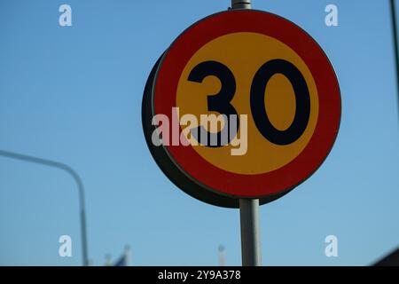 Ein hellgelbes Geschwindigkeitsbegrenzungsschild mit 30 km/h steht hoch vor einem klaren blauen Himmel und führt die Fahrer bei Tageslicht auf einer stark befahrenen Straße Stockfoto