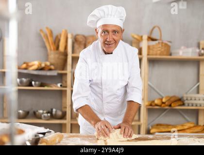 Senior Mann arbeitet in der Bäckerei als Bäcker, Knete Teig, arbeitet mit Mehl. Stockfoto