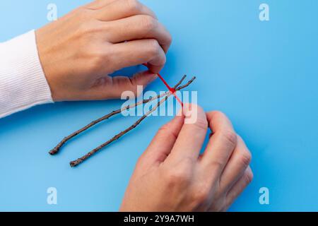 Hände, die rotes Garn am Ende an zwei Zweigen zusammenbinden, aus Stäbchen hergestellte Bastelarbeiten, Bauarbeiten für Weihnachten, Stockfoto