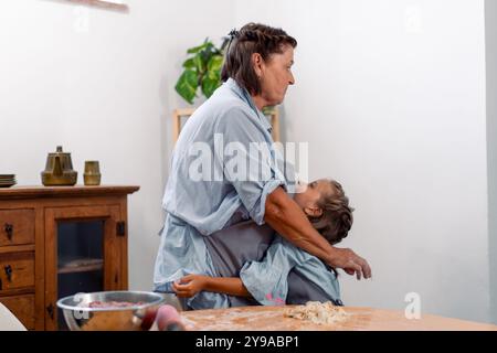 Die liebevolle Großmutter umarmt Enkelin, während sie am Tisch steht und Teig für das Gebäck zubereitete. Das liebevolle Mädchen sieht eine ältere Frau an, ein Backkonzept Stockfoto