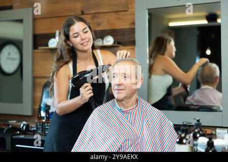 Der Friseur für Mädchen vervollständigt die Erstellung von Friseuren und trocknet die Haare mit einem Haarfön für ältere männliche Kunden Stockfoto