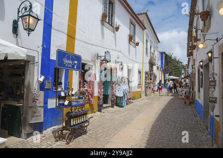 Enge Gasse mit Souvenirläden und farbenfroher Kleidung, blauen und weißen Fassaden und Blumen, R. Direita, Obidos, Obidos, Oeste, Centro, Estremadura, P Stockfoto