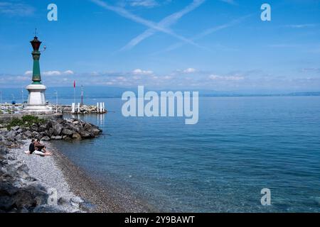 Mann, der am Ufer des Lemansees mit dem Leuchtturm im Hintergrund in Evian les Bains in den Alpen im Departement Haute-Savoie entspannt Stockfoto