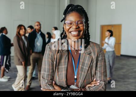 Selbstbewusste Geschäftsfrau, lacht und steht während einer Konferenz in einer Büroumgebung, während Kollegen im Hintergrund Gespräche führen Stockfoto