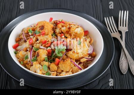 Gebratener Blumenkohl-Kichererbsen-Quinoa-Salat mit Walnüssen, roter Paprika, Zwiebeln und Koriander in weißer Schüssel auf schwarzem Holztisch, Nahaufnahme Stockfoto