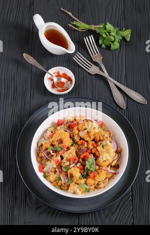 Gebratener Blumenkohl-Quinoa-Salat mit Walnüssen, rotem Pfeffer, Zwiebeln und Koriander in weißer Schüssel auf schwarzem Holztisch mit Gabeln, vertikale Ansicht Stockfoto