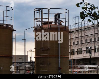 Große braune Industrietanks mit Metallleitern stehen hoch vor einem bewölkten Himmel. Dahinter befindet sich ein Gebäude mit freiliegenden Stahlrahmen und Gerüsten Stockfoto