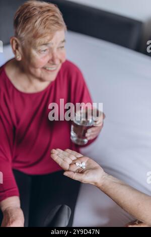 Eine ältere Frau sitzt auf ihrem Bett und ein Freiwilliger gibt ihr Pillen und ein Glas Wasser. Hochwertige Fotos Stockfoto