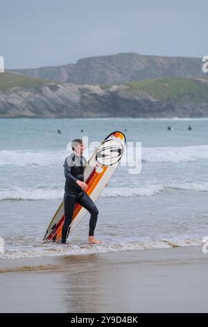 Ein glücklicher männlicher Surfer, der sein Surfbrett trägt und am Towan Beach in Newquay in Cornwall in Großbritannien aus dem Meer wandert. Stockfoto