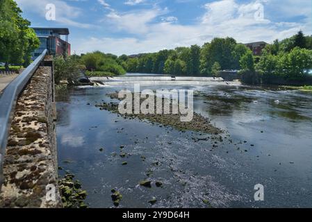 Das Foto zeigt den Fluss Ayr, der durch die Stadt Ayr fließt. Schöner klarer, sonniger Tag. Blick nach Osten flussaufwärts, vorbei am Ayrshire College Wehr und Fischleiter. Ta Stockfoto