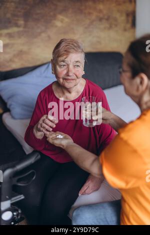 Eine ältere Frau sitzt auf ihrem Bett und ein Freiwilliger gibt ihr Pillen und ein Glas Wasser. Hochwertige Fotos Stockfoto