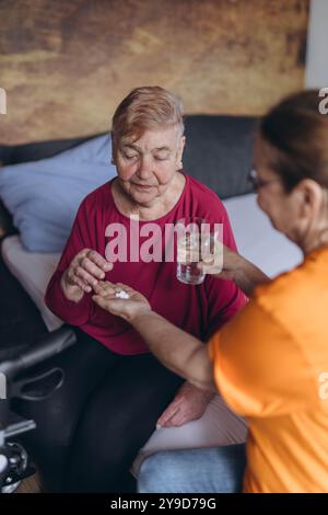 Eine ältere Frau sitzt auf ihrem Bett und ein Freiwilliger gibt ihr Pillen und ein Glas Wasser. Hochwertige Fotos Stockfoto