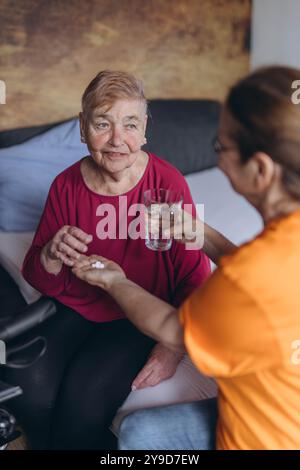 Eine ältere Frau sitzt auf ihrem Bett und ein Freiwilliger gibt ihr Pillen und ein Glas Wasser. Hochwertige Fotos Stockfoto
