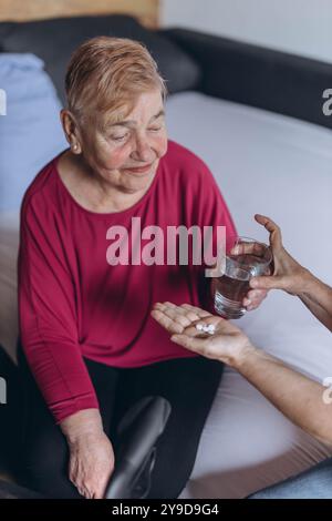 Eine ältere Frau sitzt auf ihrem Bett und ein Freiwilliger gibt ihr Pillen und ein Glas Wasser. Hochwertige Fotos Stockfoto