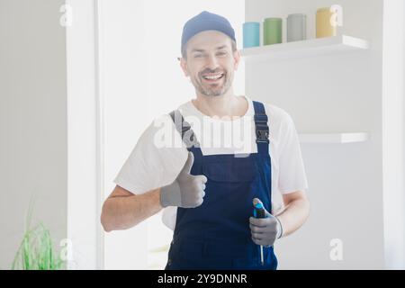 Ein lächelnder Handwerker in Overalls hält sich während der Renovierung des Hauses mit farbenfrohen Farbkannen die Daumen hoch Stockfoto