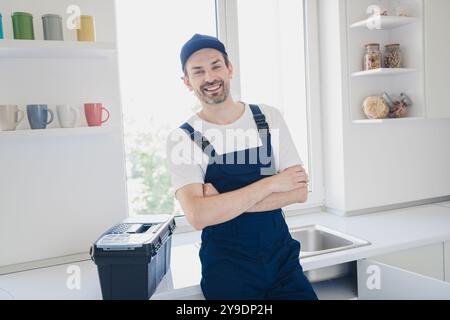 Lächelnder Handwerker in blauen Overalls, der selbstbewusst in einer hellen Küche steht und einen Werkzeugkasten für Renovierungsarbeiten zu Hause hat Stockfoto