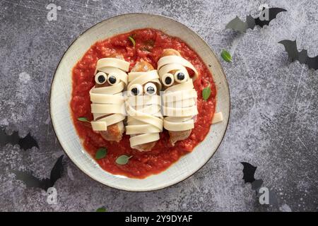 Drei Hot Dogs mit gezeichneten Augen sitzen auf einem Teller Spaghetti mit roter Sauce. Der Teller ist mit einem gruseligen Halloween-Thema dekoriert, Addin Stockfoto