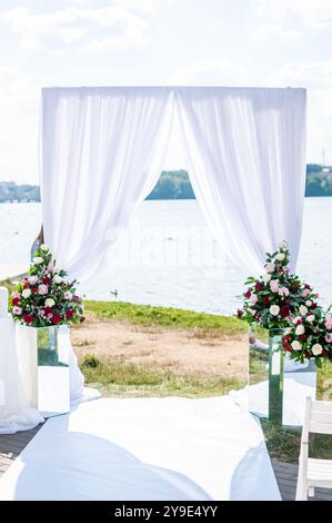 Elegante Hochzeitseinrichtung im Freien am Wasser mit Blumenarrangements und drapiertem Baldachin. Stockfoto