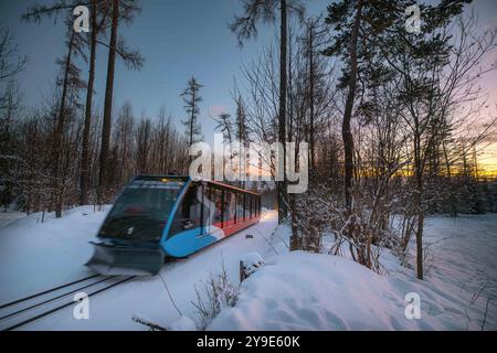 Eine abendliche Winterabfahrt: Eine elegante Standseilbahn gleitet durch einen schneebedeckten Wald, während die Sonne untergeht und ein warmes Licht über die ruhige Landschaft wirft. Stockfoto