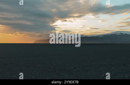 Ein atemberaubender Sonnenuntergang wirft warme Farbtöne über den schwarzen Sand einer isländischen Landschaft, mit fernen Bergen und einem stimmungsvollen Himmel, die eine ruhige Szene schaffen Stockfoto