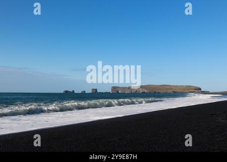 Ein ruhiger schwarzer Sandstrand trifft auf die sanften Wellen des Ozeans unter einem klaren blauen Himmel mit entfernten Felsformationen, die die malerische Landschaft ergänzen Stockfoto