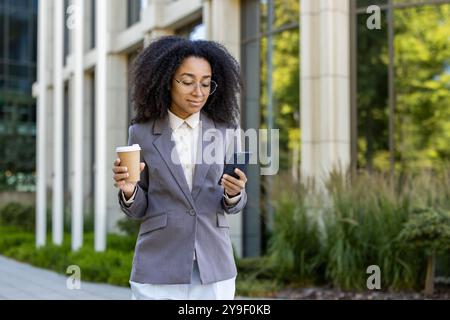 Selbstbewusste afroamerikanische Geschäftsfrau im grauen Anzug, die ihr Smartphone überprüft, während sie im Freien eine Kaffeetasse hält. Das Bild erfasst modernen urbanen Lebensstil, professionelle Kleidung und mobile Technologie. Stockfoto
