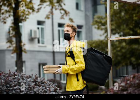 Ein Liefermann trägt eine schwarze medizinische Maske und trägt an einem regnerischen Tag Pakete in ein Bürogebäude. Stockfoto