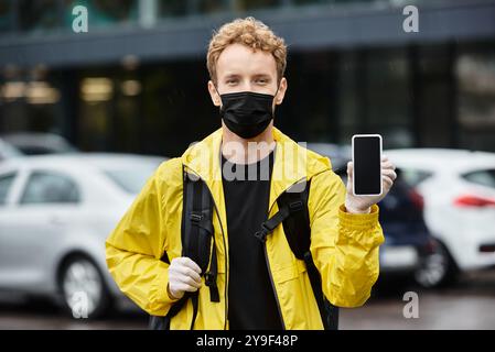 Ein Liefermann mit schwarzer medizinischer Maske bringt einen Auftrag in ein Bürogebäude, während er ein Telefon trägt. Stockfoto