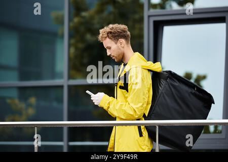 Ein Liefermann in einer gelben Regenjacke überprüft sein Handy, während er eine Bestellung in ein Bürogebäude bringt. Stockfoto