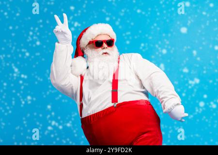 Foto von coolen funky übergewichtigen weihnachtsmann mit großen Bauch Bart Tanz Weihnachten Diskothek tragen Kopfbedeckung Hosenträger Overalls Sonnenglas Isoliert Stockfoto