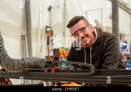 Instandhaltungstechniker reparieren eine numerische Steuerung zum Schneiden von Holz mit einem Schraubenschlüssel in der Hand, einer Schutzbrille, einem schwarzen Sweatshirt und einem orangefarbenen Handschuh Stockfoto