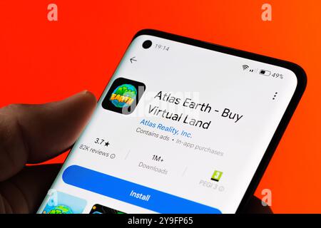 Nahaufnahme einer Hand, die ein Smartphone hält und den Installationsbildschirm der Atlas Earth App anzeigt, um virtuelles Land zu kaufen. Orangefarbener Hintergrund. Stafford, Unite Stockfoto