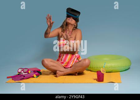 Porträt der schönen brünetten Latina Frau mit langen Haaren, die eine Virtual-Reality-Brille trägt, auf einem gelben Handtuch sitzt, rot-gelb gestreifte BI trägt Stockfoto