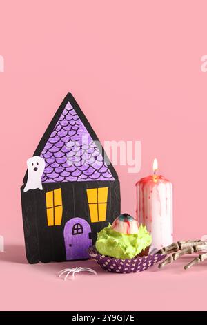 Komposition mit köstlichem Halloween-Cupcake, Papierhaus, brennender Kerze und Skeletthand auf pinkfarbenem Hintergrund Stockfoto